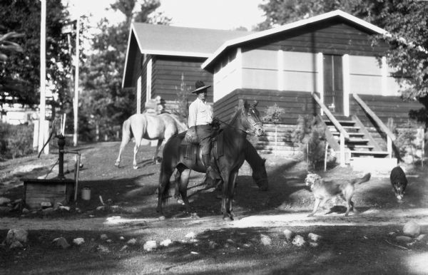 View up hill of a man sitting on a horse in front of cabin with a log foundation. Two other unsaddled horses are also in the yard along with two dogs. There is a hand-pump on the left.
