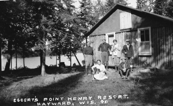Four men and two women outside lakeside cabin built with vertical logs  at Spider Lake. One women is sitting on the ground hugging a dog, and the other woman is standing holding a cat. All five people standing are wearing knee-high boots with their trousers tucked into them.