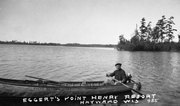Man with oar sitting in rowboat with a motor attached, paddling near shore of Spider Lake. "Point Henry Resort" is stenciled on the side of the boat.