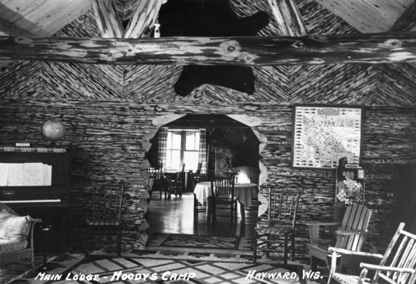 Interior view of the parlor and dining room at the lodge at Moody's Camp on Spider Lake. There are numerous chairs, an upright piano with a world globe sitting on top, a bear rug hanging on the wall, and a poster for identifying fish. The walls are logs that have been chipped. A small part of the dining room with table and chairs can be seen through the arched doorway in the log wall.