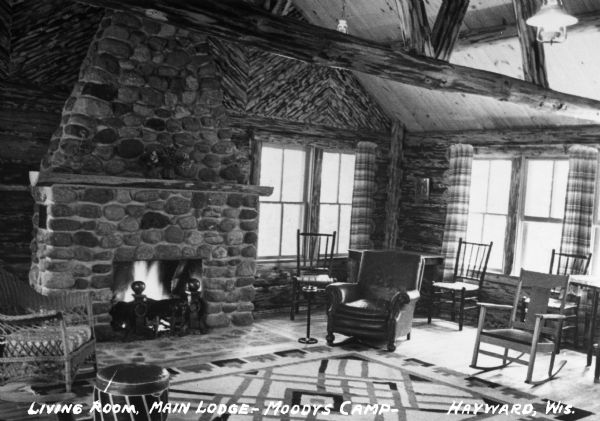 A large stone fireplace with a roaring fire is on one wall of the living room. The room has log walls and exposed ceiling beams. A variety of chairs, including a wicker rocker and a leather easy chair, are arranged around the room.