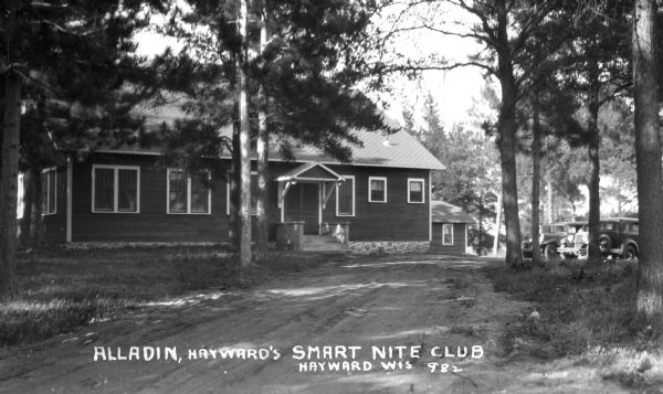View down dirt road of wood sided one-story building surrounded by tall pine trees. Two automobiles are parked nearby.