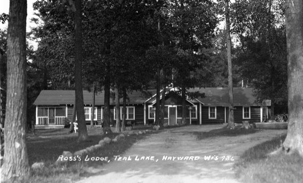 View down drive of facade of one-story long log building surrounded by trees. A man is walking along the side of the lodge in the far right background.