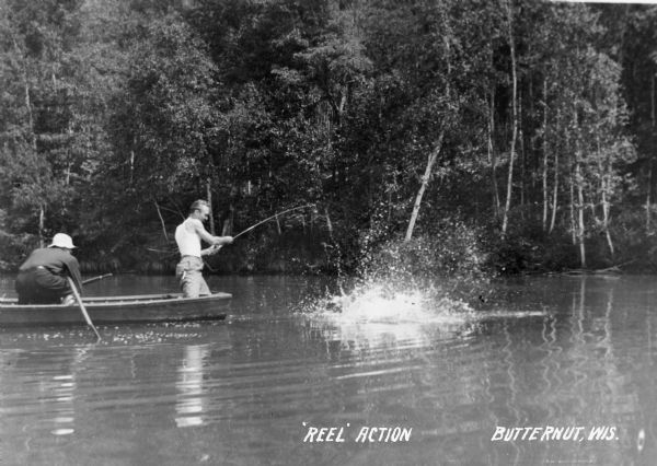 Two men, one standing one crouching, in a wooden rowboat on Forest Lake. The man standing is reeling in a fish that is making a big splash in the water.