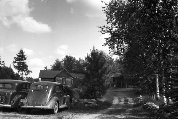 Two automobiles parked up against a stone wall in front of a log cabin near Moose Lake.