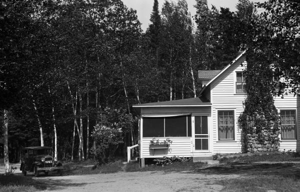 An automobile is driving up the drive towards a two-story white wood-sided building with flower planters under the windows and a field stone chimney on the outside wall.