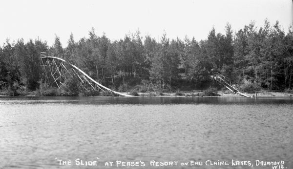 A large slide at Pease's Resort on the shore of the Upper Eau Claire Lake. Children are in the water near stairs that go up the hill.