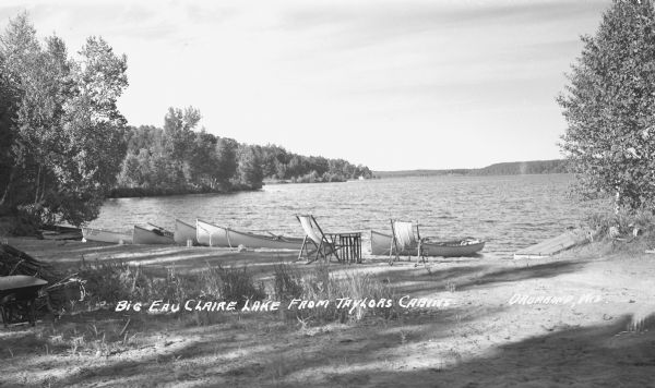 Canoes and lawn chairs at the shore of Upper Eau Claire Lake. Caption reads: "Big Eau Claire Lake From Taylors Cabins, Drummond, Wis."
