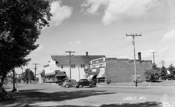 View across street of automobiles parked in front of a hardware store and a drugstore on Main Street.