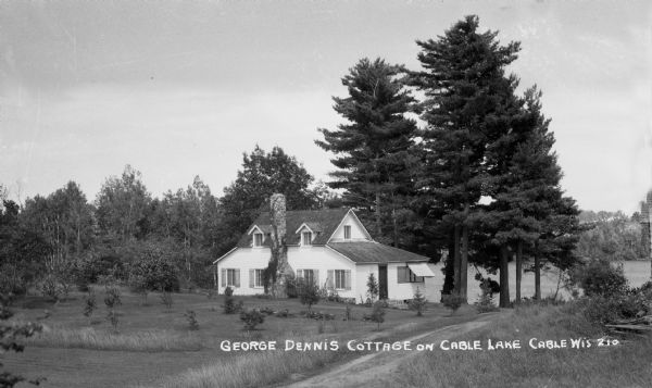 View across driveway of one-and-a-half story house with a stone chimney situated under large pine trees and new plantings. Cable Lake is in the background.
