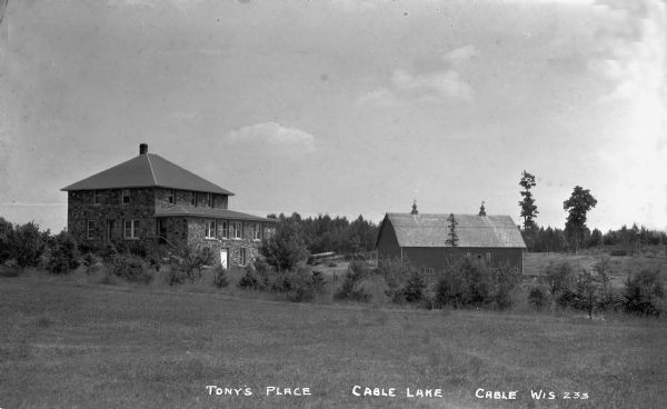 View across field of a two-story stone house with a wood barn near Cable Lake.
