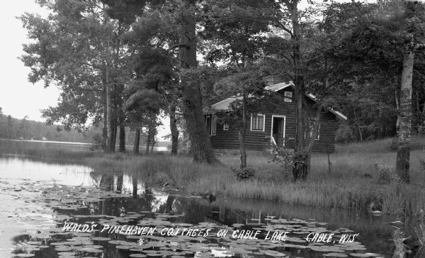 View across water filled with lily pads of a one-story cottage on shore of Cable Lake.