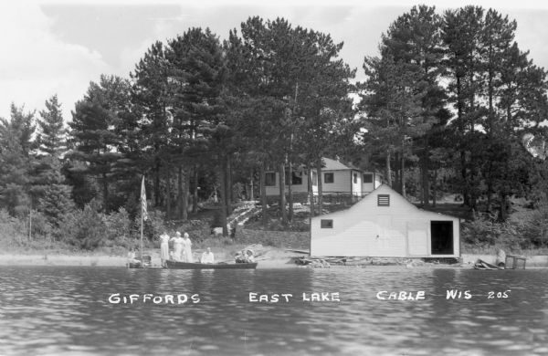 View from water of people sitting in a canoe next to a boat dock where more people are standing. A boathouse is on shore and a cottage is up a slight hill in the background.