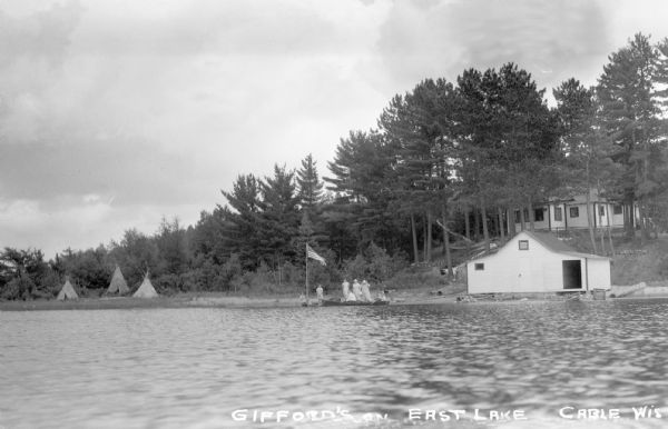 View from water of people sitting in a canoe on the shore of East Lake next to a dock on which people are standing and a flag is waving. On shore there is a boathouse to the right and tepees to the left. There is a cottage in the background.