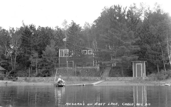 View from water of a boy sitting on the back of a wooden boat with his feet on a pier. He is holding a dog which is standing on the dock. There is a small outbuilding along the shore and a cottage with a stone retaining wall up the hill.