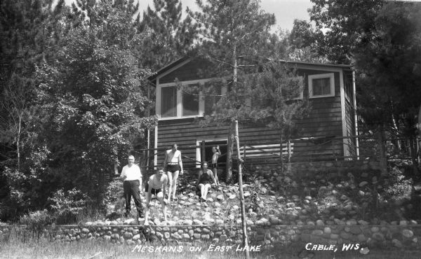 View from shoreline of family members dressed in bathing suits, sitting on rock strewn incline in front of a cottage. One man is dressed in trousers and shirt, smoking a cigar.
