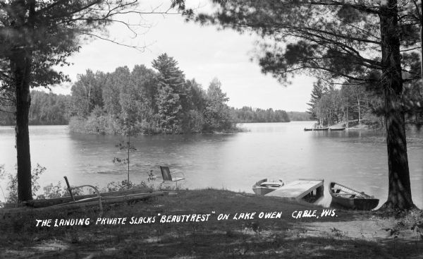 Point of land in lake with lawn chairs in the shade of trees and wooden boats tied to boat dock on Lake Owen. Caption reads: "The Landing at Private Slack's 'Beautyrest' on Lake Owen, Cable, Wis."