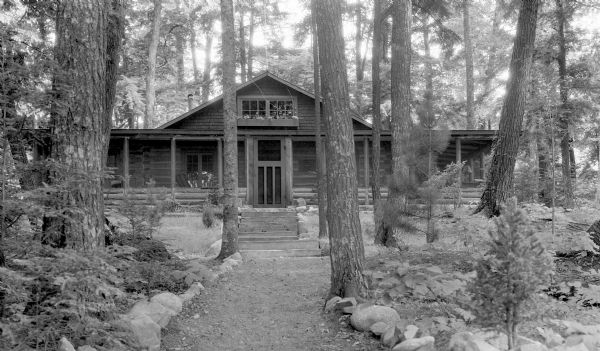 Stone-lined path through woods leading to a one-story log cabin with a wrap-around porch and window boxes with flowers. The cabin is located near Cable.