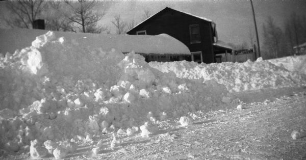 Winter scene with snow plowed high on County Road M, Clam Lake (Ashland Co.), Wisconsin. The Chippewa Tavern is visible in the background, and the building in the foreground has snow plowed to the eaves.