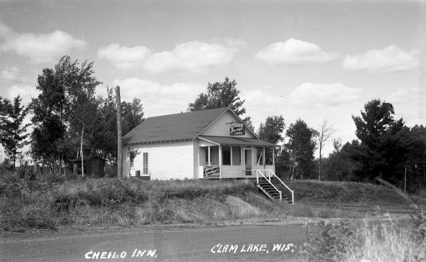 View from across road of a one-story building with stairs leading from the dirt road to the porch; an outhouse is behind the inn. Numerous beer signs are on the buildings, including "Leinenkugel's, Breunigs, and Grain Belt". Cheilo Inn was near Clam Lake.