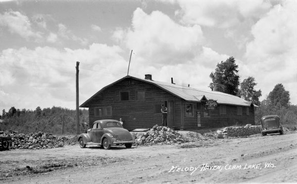 A man is exiting a wooden building with a Blatz beer sign hanging on the front and two automobiles parked nearby. Near Clam Lake.
