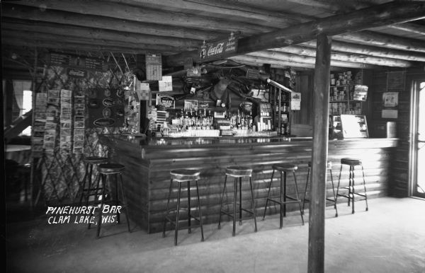 Interior of a tavern near Clam Lake. Bar stools are around a bar, and the walls covered with animal mounts, guns, traps, postcards, and signs. Liquor and canned goods are behind the bar.