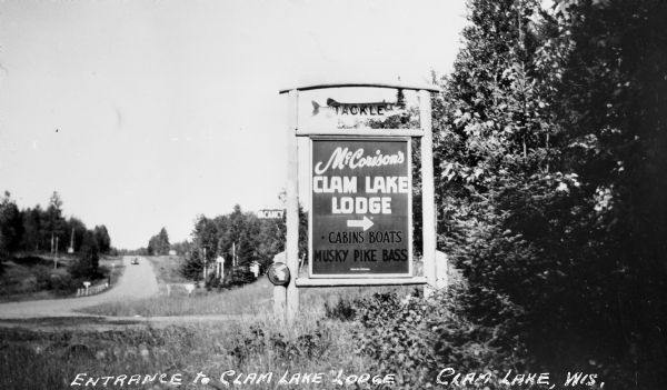 A car can be seen in the distance driving down the highway towards an entrance sign for "McCorison's Clam Lake Lodge-Cabins Boats Musky Pike Bass."