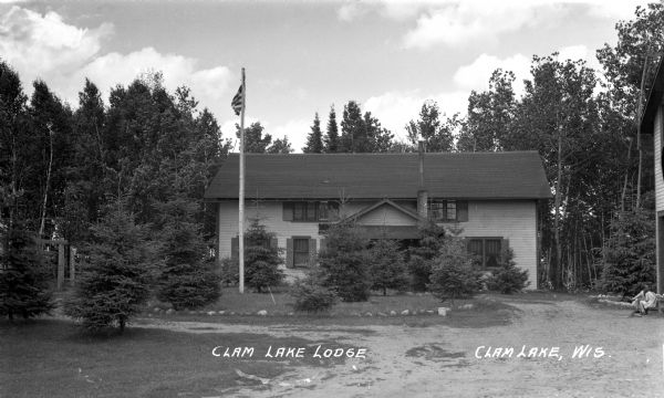 Exterior view of two-story wood building with a flagpole surrounded by young spruce trees. Two children are sitting on the steps of a building to the right.
