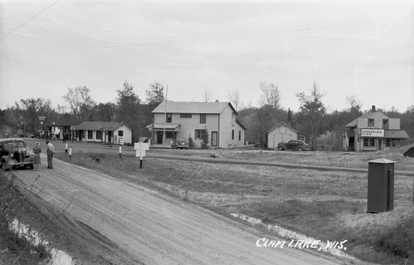 Elevated view across road of people and automobiles in front of homes and businesses along a dirt road. In the background is a local grocery store, tavern and service station. Caption at bottom right reads: "Clam Lake, Wis."