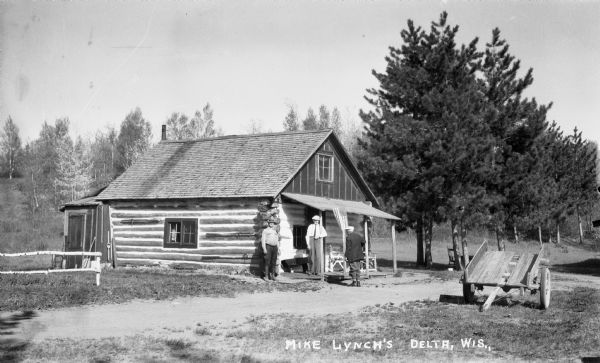 Exterior view of three men standing in front of a log cabin. A wagon sits nearby. A flag hangs off the porch, and oars are stored on the exterior cabin wall.