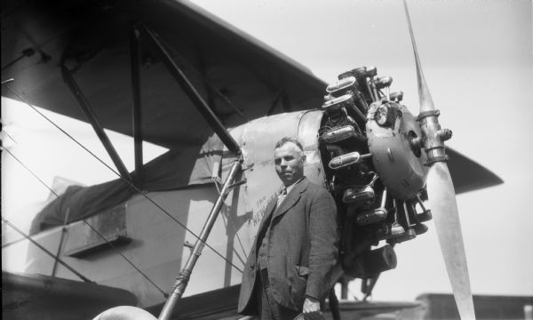 A man wearing a suit and holding his hat is standing near the front of a single prop airplane.