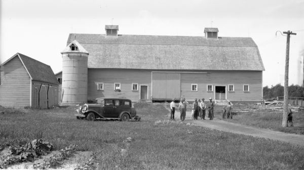 Group of men and a child standing near a barn and silo. A car is parked nearby.