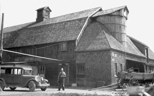 A farmer is standing next to an automobile in front of a barn.