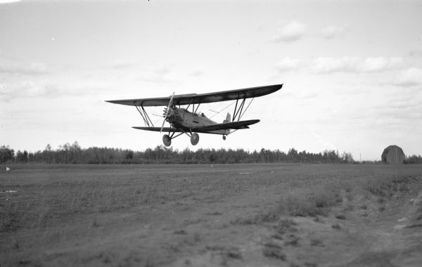A biplane is flying low over a field with a barn in the background.
