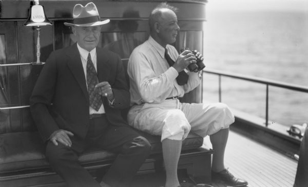Two men sitting on board cabin cruiser "Lamora" on Lake Superior. On the left is a bell with the name of the cruiser engraved on it.