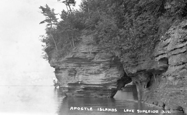View across water of trees and plants covering a sandstone cliff and arch on the shoreline along Lake Superior. Another sandstone arch is in the far background.