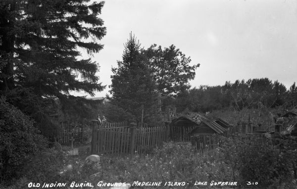 Ojibwe wooden burial houses surrounded by wooden fence on Madeline Island. Two women stand in the background.