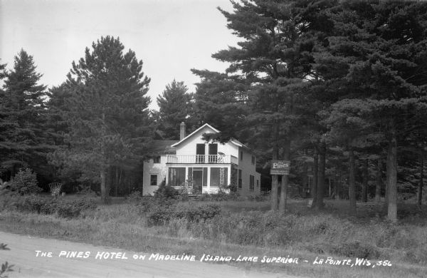 View from road of two-story white hotel with a screen porch; a sign advertising "The Pines" is attached onto two pine trees in front of the hotel.