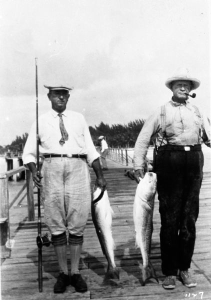Two men standing on dock each holding a lake trout.