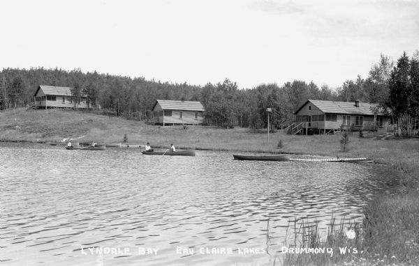 View from water of men fishing from boats along lakeshore in front of three cabins on Upper Eau Claire Lake.