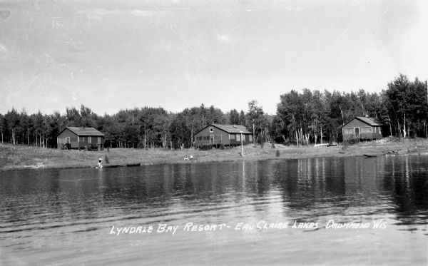 View from water of people along the shoreline of the Upper Eau Claire Lake. In the background are trees and three cabins, each with an automobile parked nearby.