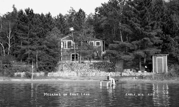 View from water of a woman wearing a bathing suit sitting on the dock in front of a cabin. Five people stand on the porch of the cabin looking out over the lake. There is another small building near the shoreline.