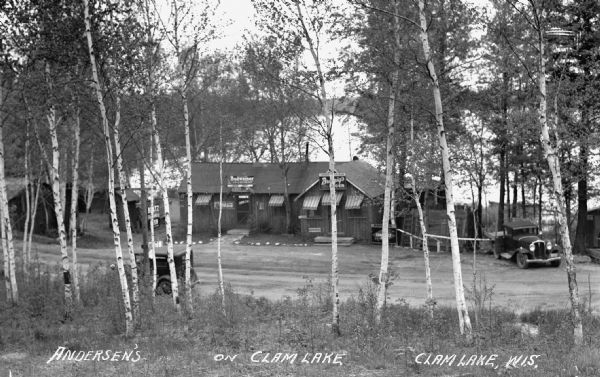 View down hill of a tavern, with signs advertising "Budweiser" and "Beer," on the shores of Clam Lake. There are numerous outbuildings and two automobiles parked along the road.