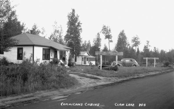 View from road of a man, woman and boy sitting in the entry way of a cabin, with another cabin, a canoe, and an automobile on the right. A sign says "Clam Lake Resort, Boats and Cabins, Cormican's Place."