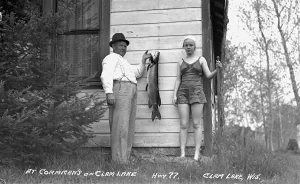 Standing outside of a cabin at Cormican's on Clam Lake is a man holding up a large fish and a girl in a bathing suit and cap holding a paddle.