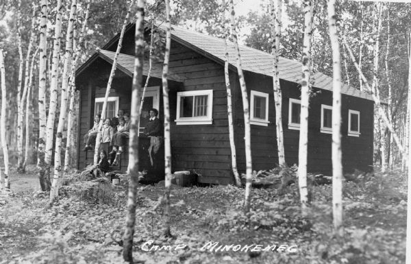 Group of two women and five girls standing and sitting on the front stoop of a cabin at Camp Minokemeg on Buffalo Lake. The cabin is surrounded by small, possibly birch, trees.