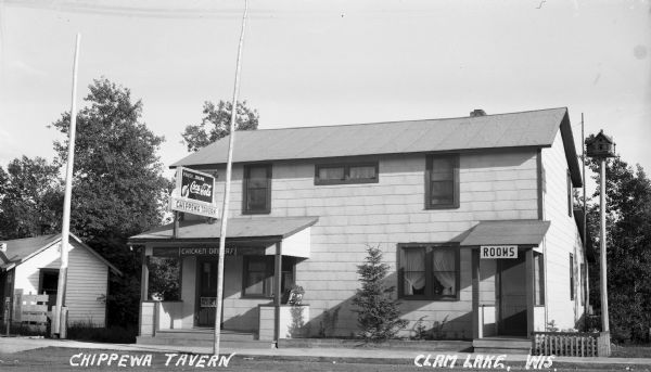Exterior view of the Chippewa Tavern, a two-story building with two main doorways. The door on the right has a sign that says "Rooms," while the door on the left has signs that read "Pause... Drink Coca-Cola," "Chippwa Tavern" and "Meals Short order, Chicken Dinners, Lunches Sandwiches."