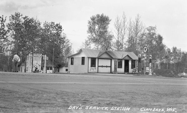 View across road of a one-story building, Day's Service Station, with gasoline pumps and signs and a map out front. The signs read “Straight Ahead, 10 Mi. then turn left 3Mi. to the North end of Namakacon Lake.” “Let us MARFAK your car,” “Good Rich Tires” and “Texaco Gasoline Motor Oil.” Two men stand talking on the far left.