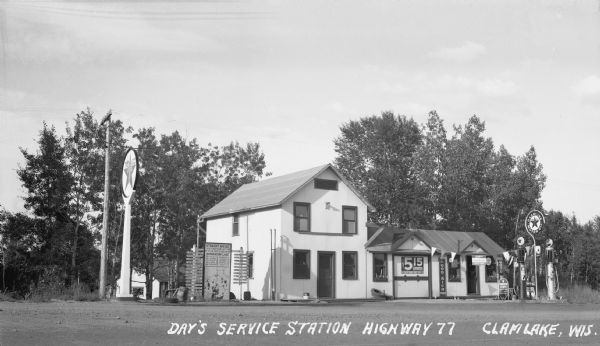 View across road of Day's Service Station with gasoline pumps, maps and signs out front. The right half of the building is one-story high, and the left half of the building is a two-story addition. The signs out front read “Straight Ahead, 10 Mi. then turn left, 3 Mi. to the North end of Namakacon Lake.” “Quaker State Motor Oil,” “GoodRich Tires” and “Texaco Gasoline Motor Oil.”