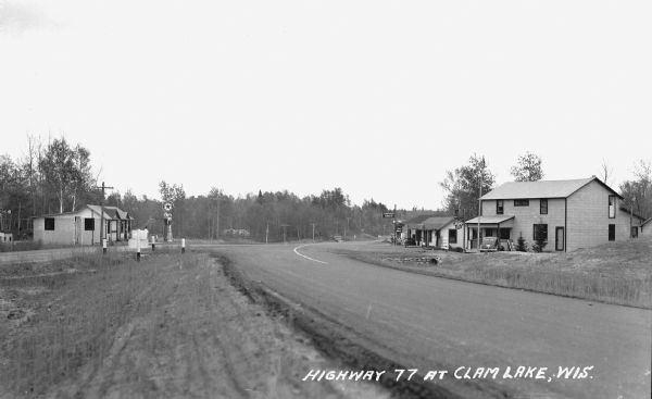 View looking down Highway 77 at Clam Lake. On the left is a one-story building, Day's Service Station. On the right are two buildings, the one-story Standard Service Station building and a two-story building with a sign for beer. Men are sitting on the front porch of the Standard Service Station. (In a 1943 photograph this two-story building is called the Chippewa Tavern.)